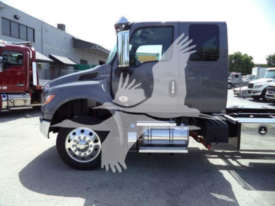 Truck side view showing a gray International MV cab with chrome fuel tank and steps, rollback bed visible, wheel front tire, side window, and dealership lot background.