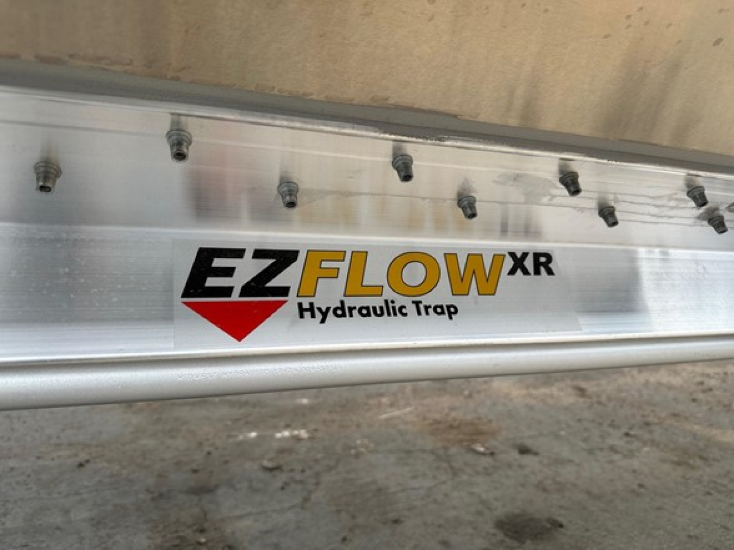 EZFLOW XR hydraulic trap sticker on aluminum trailer side rail with bolts and gravel ground in background.