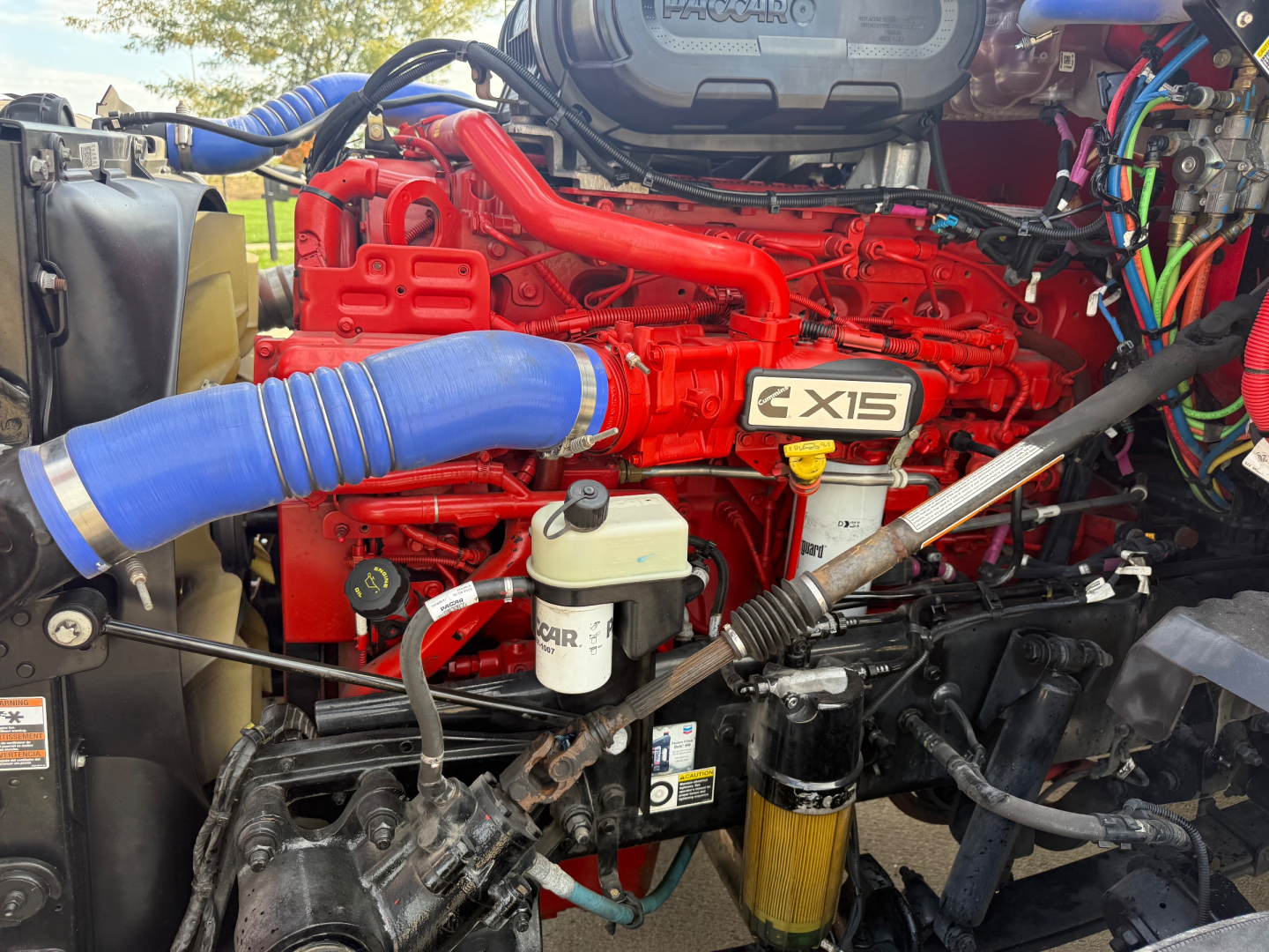 Kenworth T680 engine bay showing red engine block with X15 badge, blue intake hose, white reservoir, black hoses, and yellow oil canister.