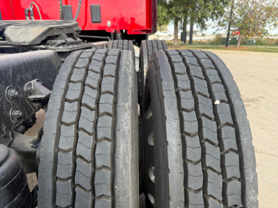 Kenworth T680 truck dual rear tires with tread pattern visible, chassis components and red cab in background.
