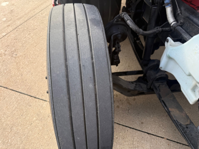 Kenworth T680 front wheel and suspension area with tire tread visible and parts of chassis and coolant reservoir.