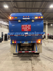 Blue Peterbilt 320 rear view in a garage with Wayne logo, red warning sign, license plate, and tow hitch.