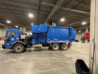 Peterbilt 320 garbage truck in a warehouse setting, blue body with attached solid waste compactor and container, side view showing cab and rear compactor unit.