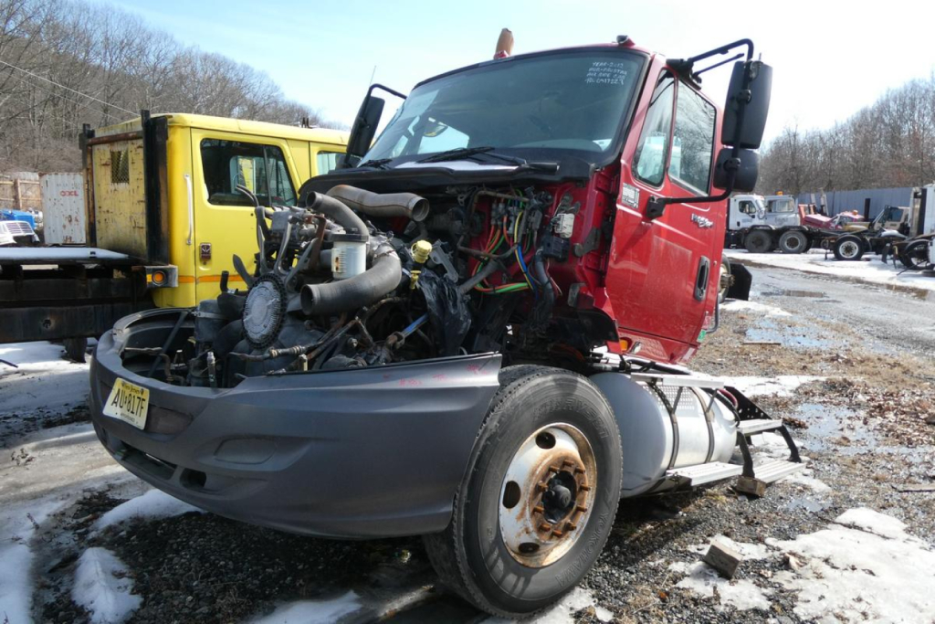 Truck engine bay exposed on red International PROSTAR cab with visible wiring and components; front bumper and wheel well in view; other trucks in background.