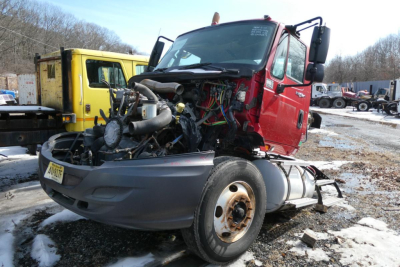 Truck engine bay exposed on red International PROSTAR cab with visible wiring and components; front bumper and wheel well in view; other trucks in background.