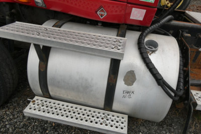 Truck fuel tank with metal steps and a black hose; faint handwritten note near tank reading 'TANK N.G.' and a circular access port; gravel ground.
