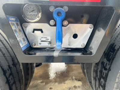 Trailer rear control panel with blue lever and gauge showing lift/tilt hardware; includes label panel with instructions and a small lever; two tires visible on either side.