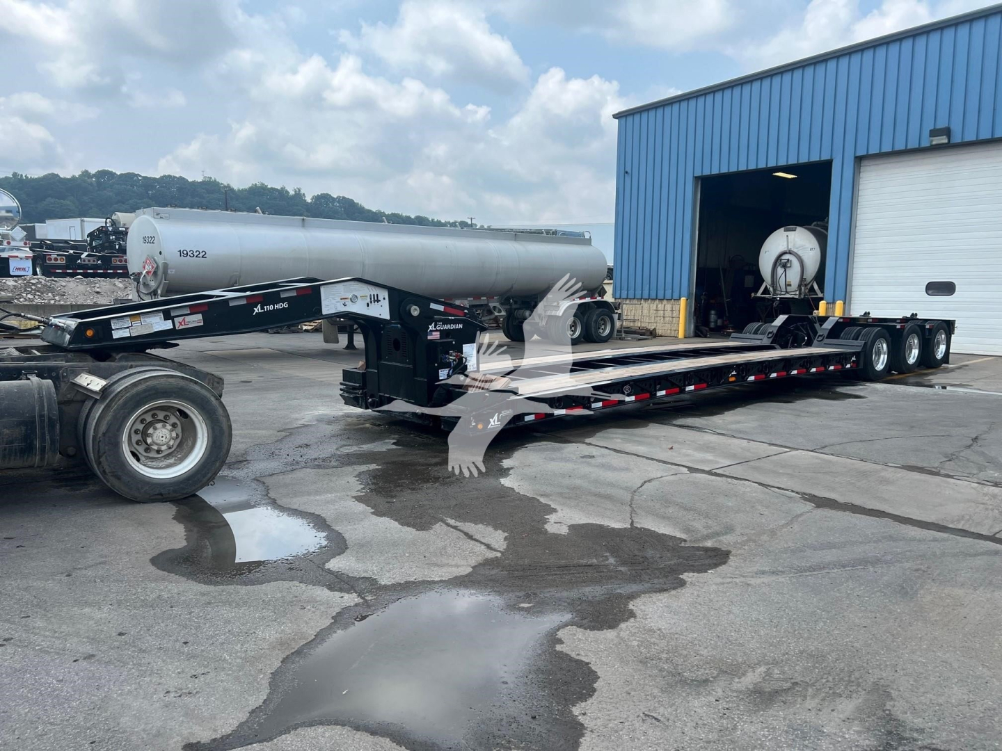 Lowboy trailer with tanker and hydraulic lines attached, extended flatbed in yard near blue warehouse.