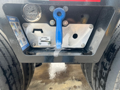 Trailer hitch control panel with blue lever and gauge on metal plate, warning decals, and dual tire tread visible at sides.