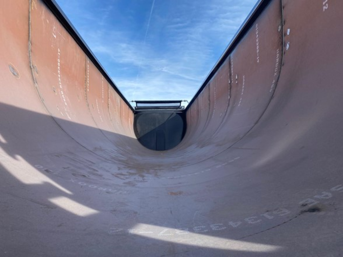 Dump trailer interior shown from the rear, curved bottom with blue sky above, wheel well opening visible at far end.