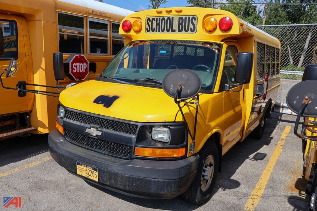 Yellow Chevrolet Express school bus with STOP sign, front view, headlights, mirrors, and ‘SCHOOL BUS’ sign on top.