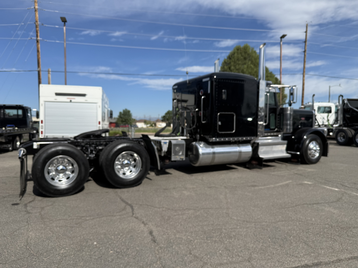Peterbilt 389 tractor-trailer in a used vehicle lot, side view showing black sleeper cab, dual rear axles, chrome fuel tanks, and white service box in background.