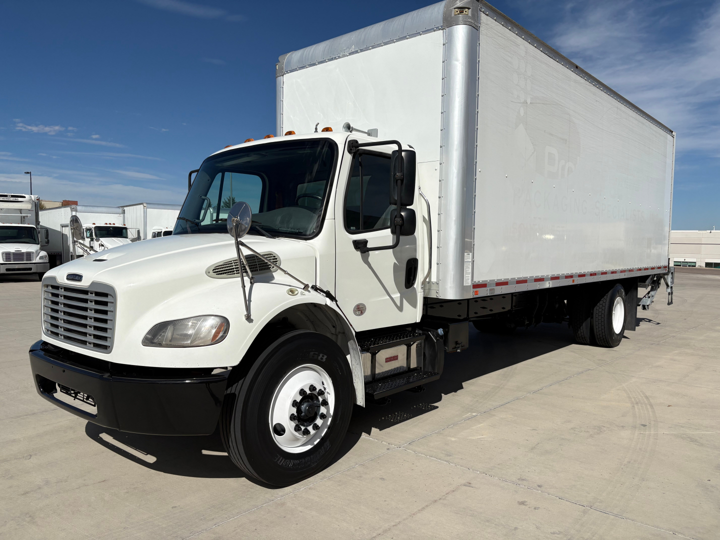 Box truck with white cab and large white box body, Freightliner M2-106. Front grille, headlights, side mirrors visible.