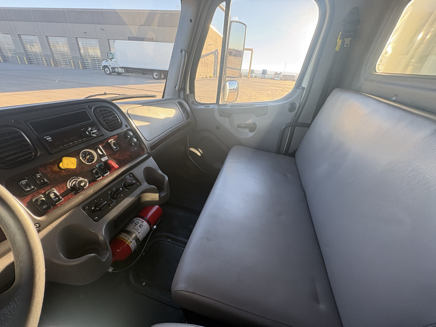 Cab interior of a 2022 Freightliner M2-106 showing dashboard with controls, steering wheel, passenger bench seat, and a fire extinguisher mounted under the dashboard.