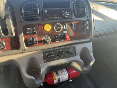 Dashboard panel of a Freightliner M2-106 cab showing gauges, switches, woodgrain trim, and a red fire extinguisher mounted under the dash.