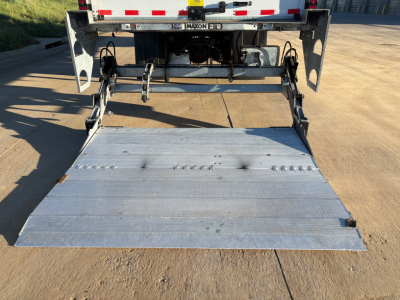 Rear view of a Freightliner M2-106 truck with a raised aluminum loading ramp and hydraulic lift system, showing the back deck and wheel wells.