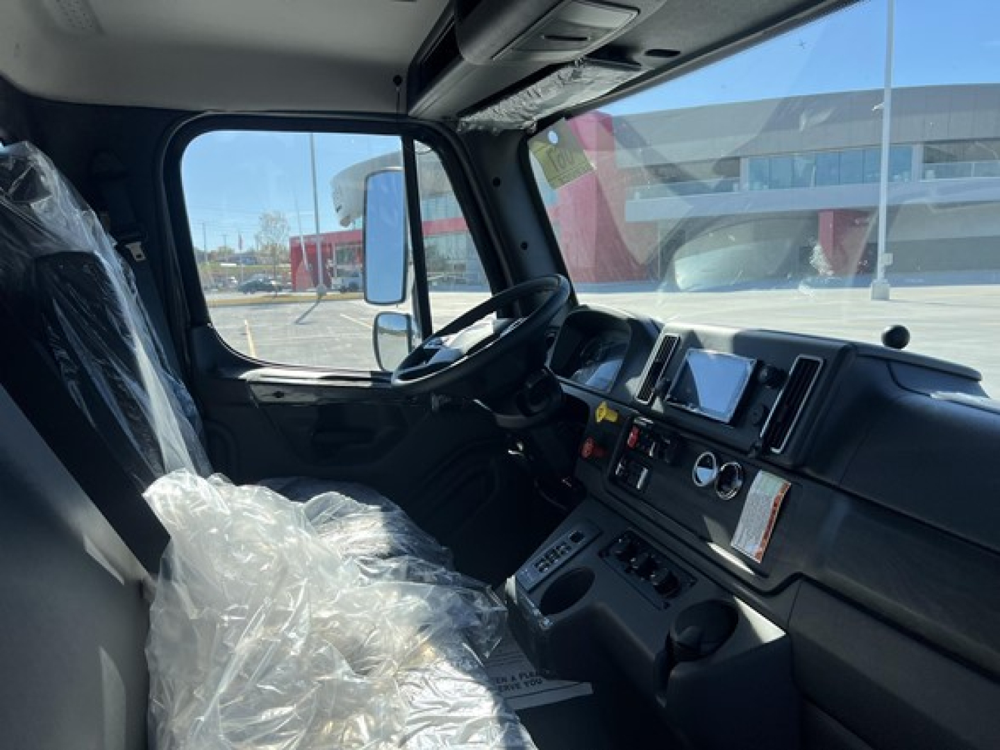 Freightliner Biz Class M2 106 cab interior view showing steering wheel, dashboard, steering column, and driver seat wrapped in plastic. Windows reveal exterior lot.