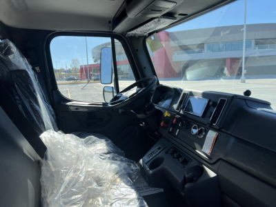 Truck cab interior of a 2026 Freightliner M2 106 showing the driver seat, steering wheel, dashboard with gauges, infotainment screen, and controls. Clear side window and exterior building reflections.
