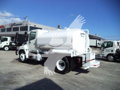 2019 Hino 338 dump truck in a used vehicle lot; side rear view shows cab and chassis with front wheels; water category listing in the scene; background includes other trucks.