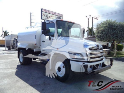 Hino 338 water tanker truck in profile view with white cab and tank, TLC Truck Sales Center sign in background.