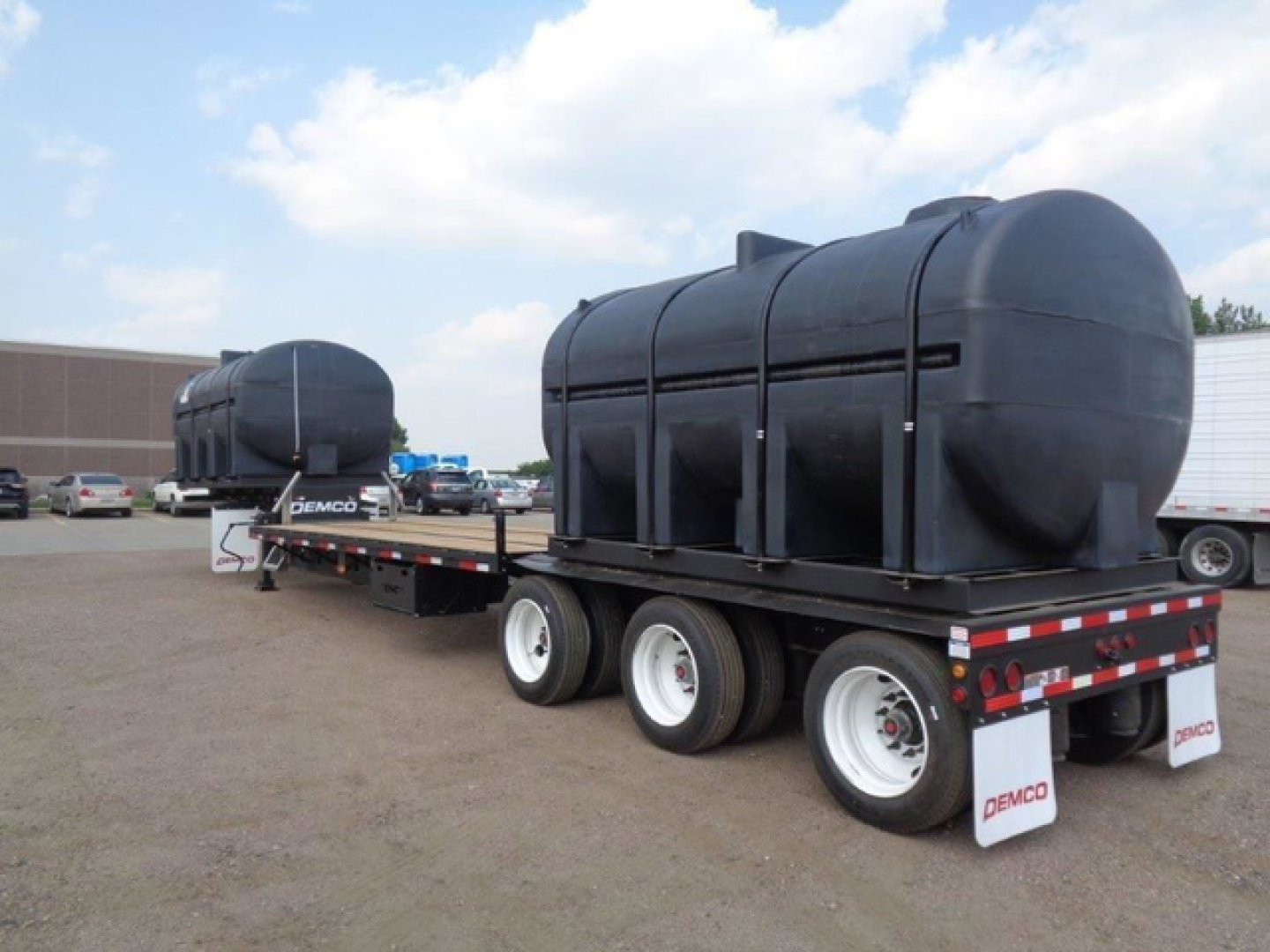 Demco Liquid Tender trailer with large cylindrical tanks mounted on a tri-axle flatbed, black tanks, white wheels, Demco branding on mud flaps, in a parking lot.
