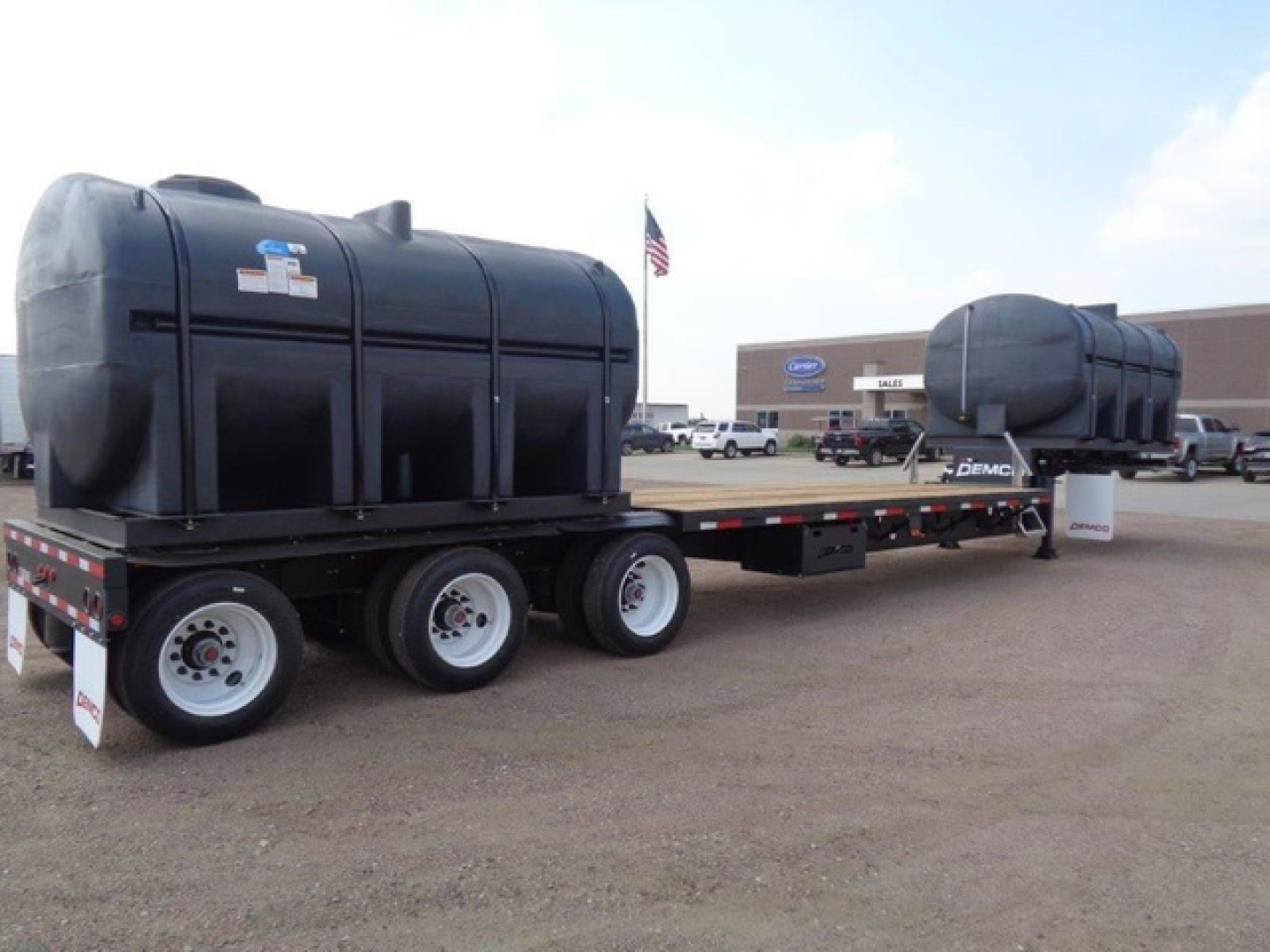 Trailer with dual large black tanks on a long flatbed trailer, three axles visible, dealership lot in background, small American flag on pole, clear sky.