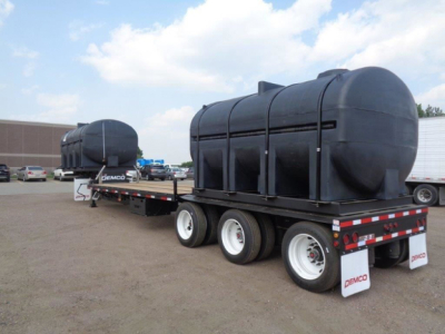 Demco Liquid Tender trailer with large cylindrical tanks mounted on a tri-axle flatbed, black tanks, white wheels, Demco branding on mud flaps, in a parking lot.