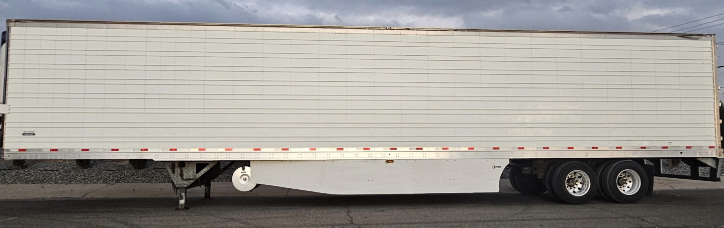 Trailer side view showing a long refrigerated trailer with tandem axles and white ribbed metal walls, no visible text.