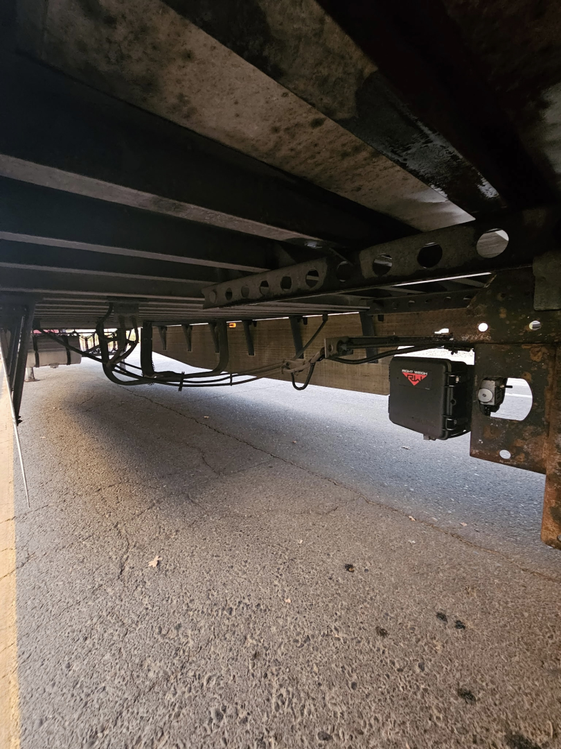 Trailer undercarriage showing wiring, frame rails, and a black electrical connection box with a red label. visible hardware and support beams.