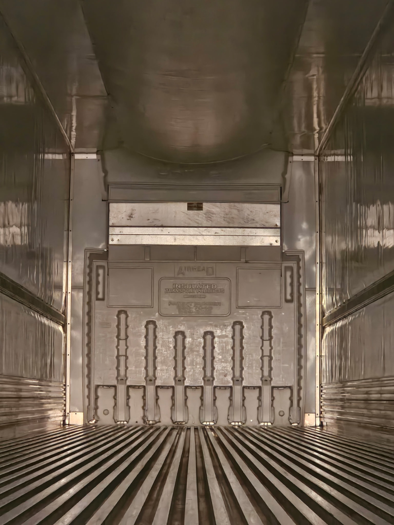 Trailer interior showing rear cargo door with vertical grooves and ribbed floor; stainless steel walls and ceiling, branding plate on door.