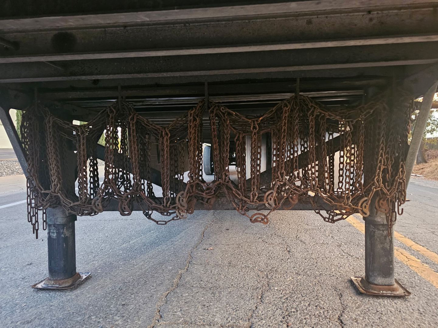 Trailer undercarriage showing multiple rust-toned chain netting and support legs, view from underneath near road surface.