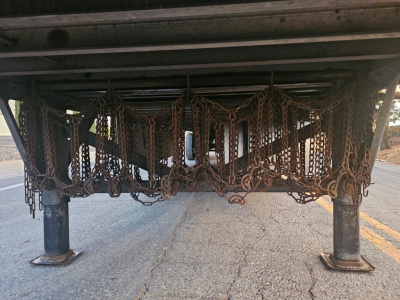 Trailer undercarriage showing multiple rust-toned chain netting and support legs, view from underneath near road surface.