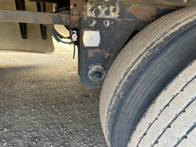 Close-up of a trailer frame near the wheel showing a small square sticker and a circular port with cables; part of a dual-axle reference frame visible.