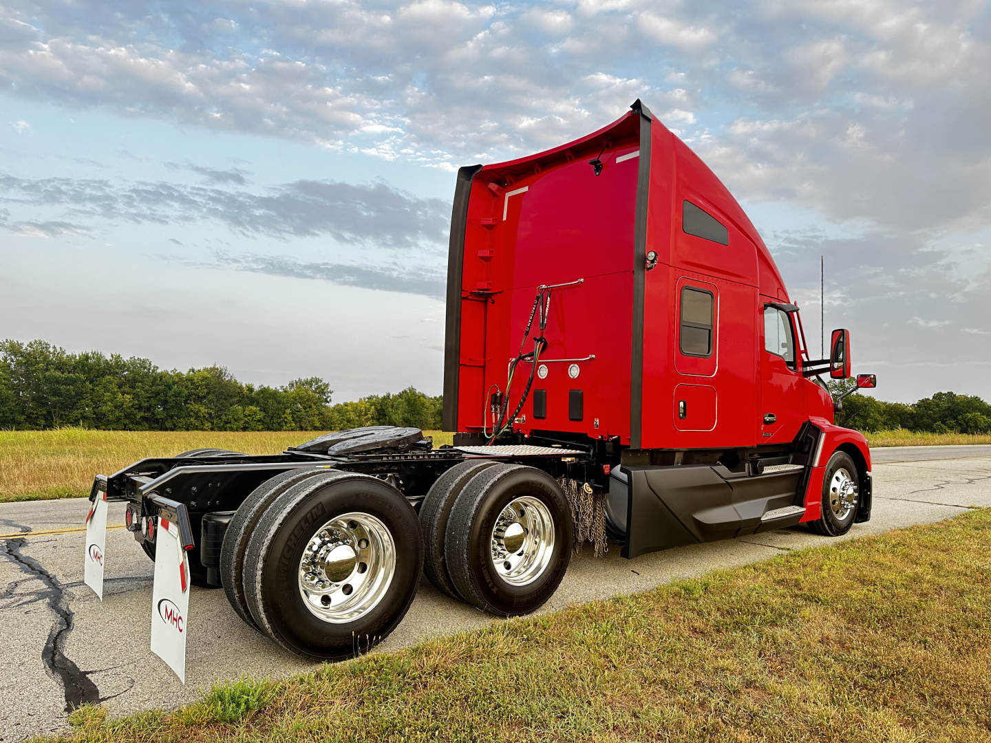 Kenworth T680 truck cab in red with sleeper, rear view showing dual rear axles and wheels, 2024 model year, on pavement with grass and cloudy sky.