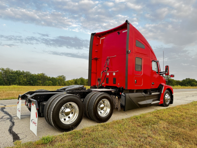 Kenworth T680 truck cab in red with sleeper, rear view showing dual rear axles and wheels, 2024 model year, on pavement with grass and cloudy sky.