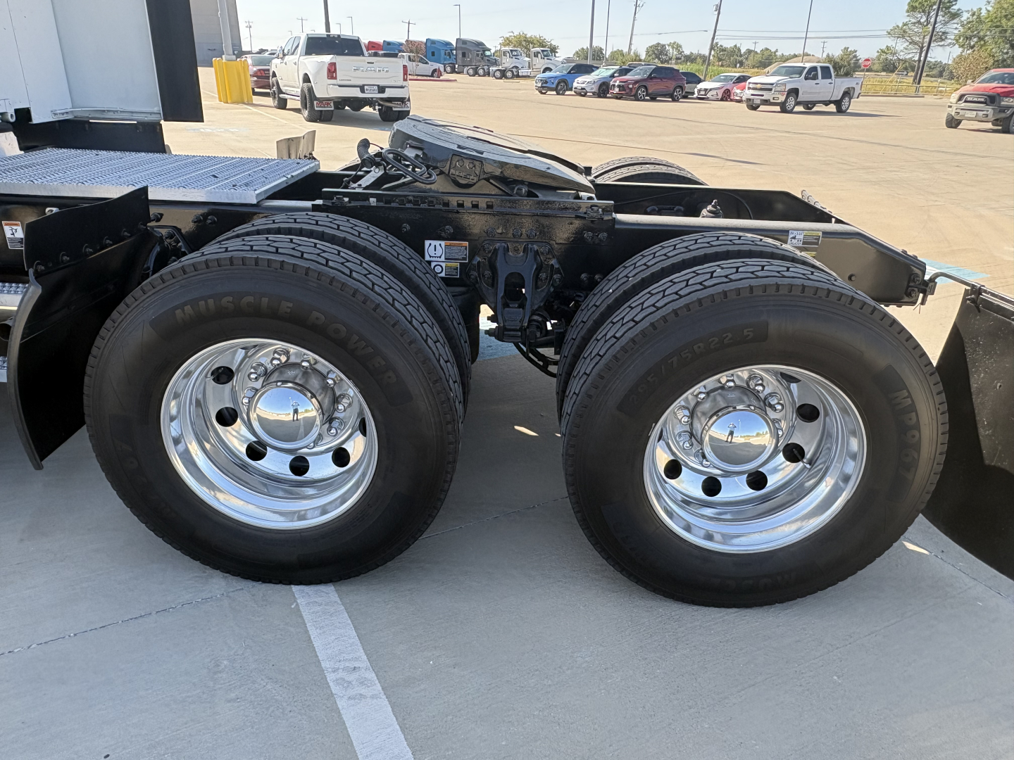 Peterbilt 579 chassis with dual rear axles and chrome wheels; visible label on frame; background shows dealership lot with other trucks.