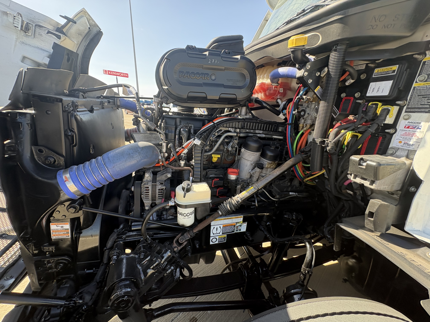 Peterbilt 579 engine bay view showing multiple components including air intake, coolant reservoir, wiring harnesses, hoses, and battery area in a sleeper cab truck.