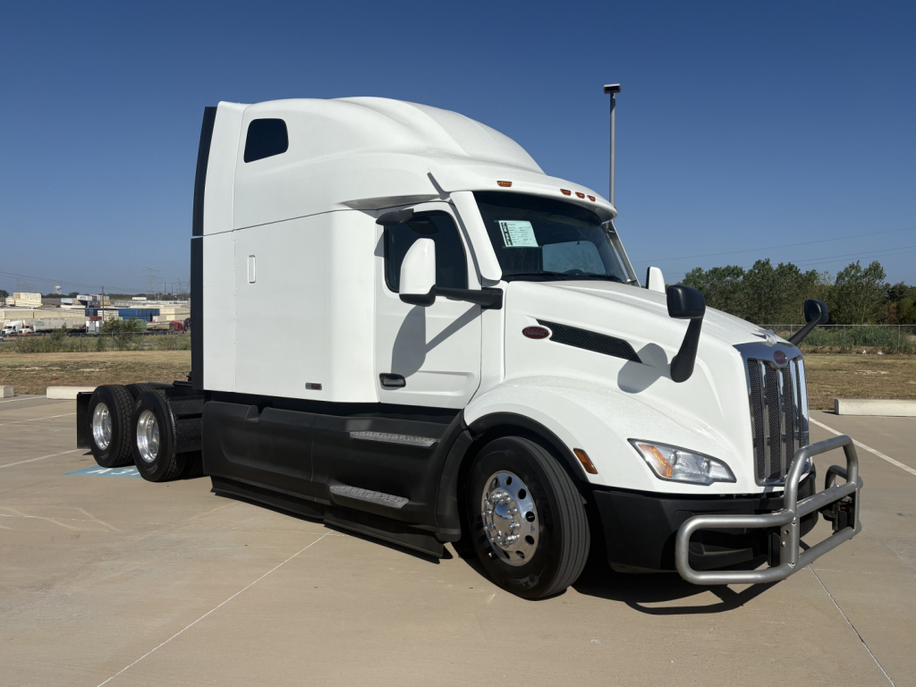 Peterbilt 579 tractor in white, side cab view, sleeper, three rear axles visible, chrome wheels, front bumper guard, clear sky.
