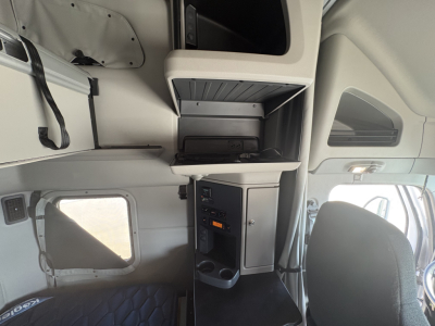 Peterbilt 579 cab interior showing sleeper area with overhead storage compartments and control panel. Visible materials are light gray, with a cup holder and a window on the left.