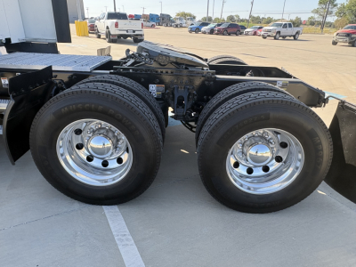 Peterbilt 579 chassis with dual rear axles and chrome wheels; visible label on frame; background shows dealership lot with other trucks.