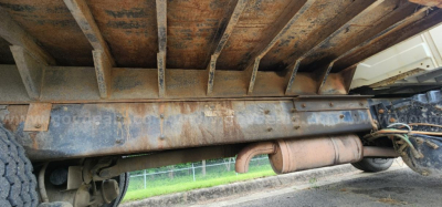 Underbody of a dump truck showing exhaust pipe and frame rail with rust-like staining and wheel in background.