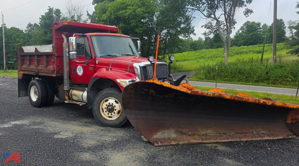 Dump truck with a large rusted front plow, red International WorkStar 7500 with black plow blade, on a road with trees in the background.