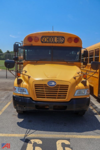 School bus front view showing yellow body, 'SCHOOL BUS' sign, blue Bird logo on grille, headlights, and mirrors in parking lot.