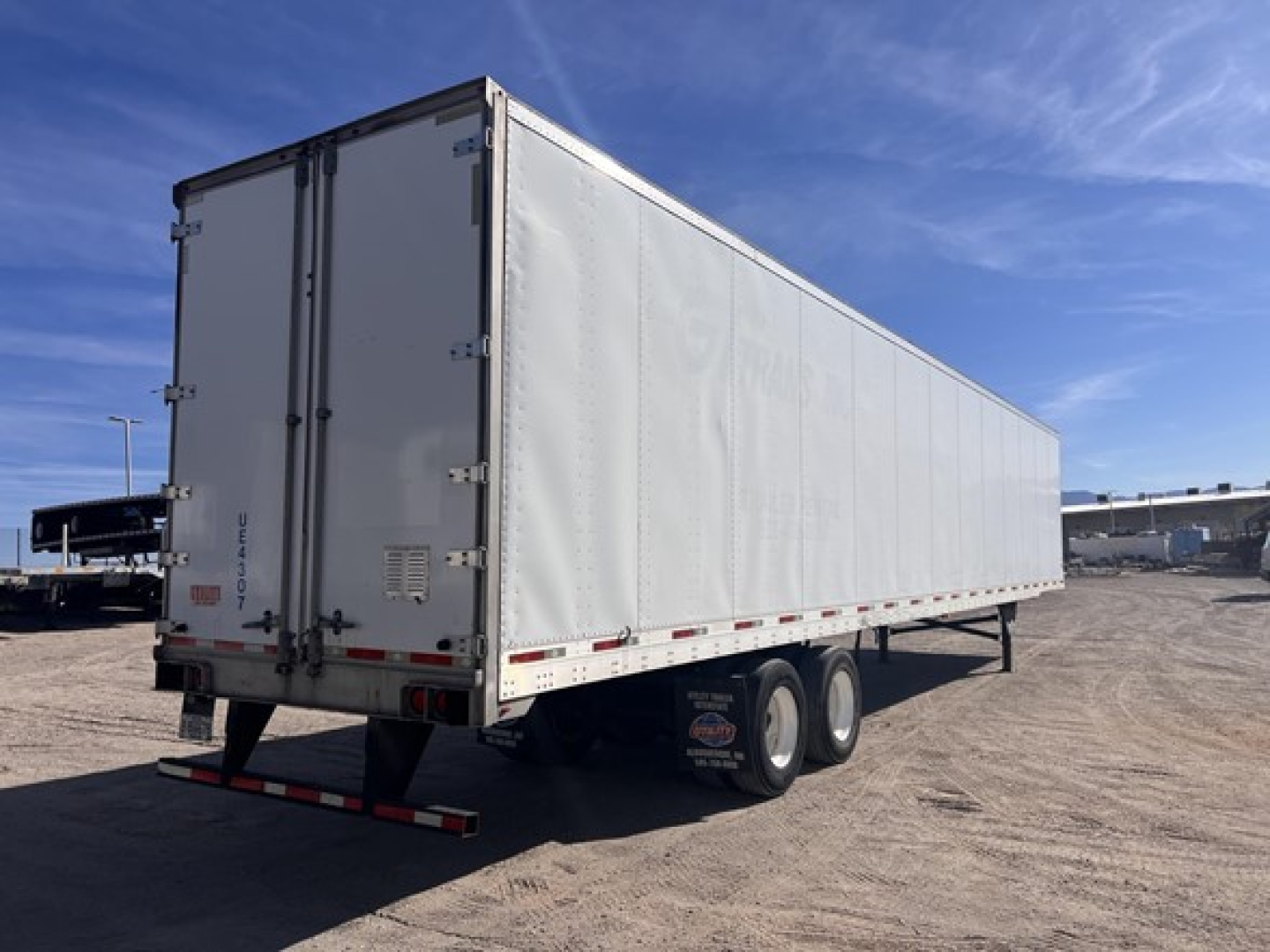 Utility trailer showing white dry van cargo area with dual rear doors, rear underride guard, and tandem axles in a gravel lot.