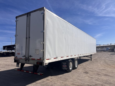 Utility trailer showing white dry van cargo area with dual rear doors, rear underride guard, and tandem axles in a gravel lot.