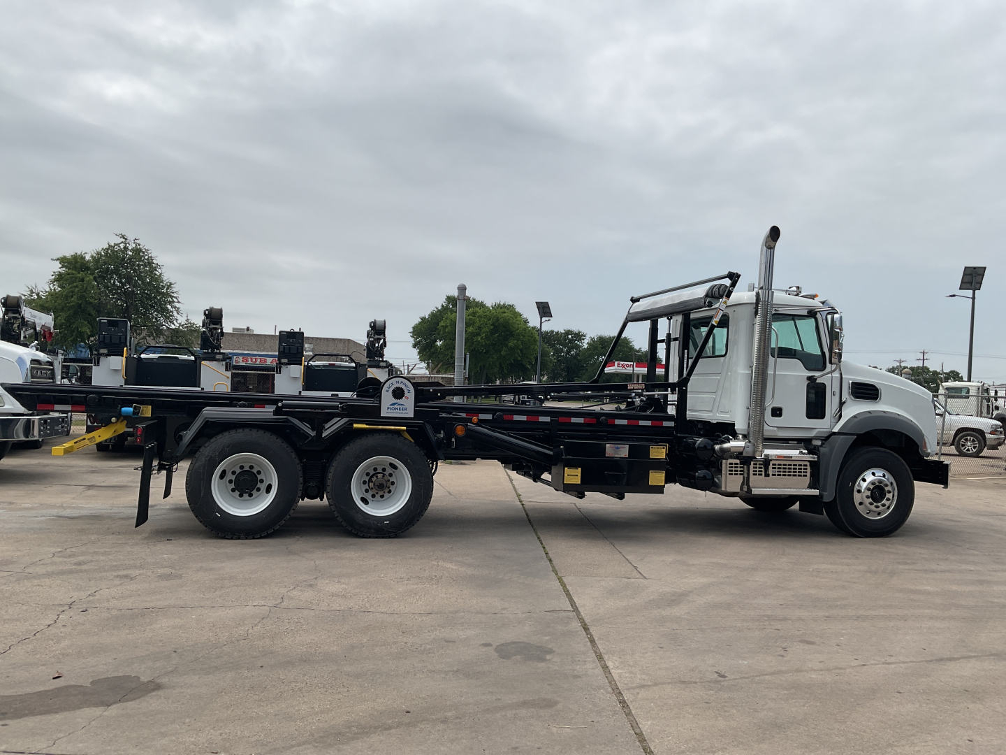 2026 Mack GR64B roll off truck side view showing cab, long chassis, and triple rear axles with a flatbed bed and hydraulic components.