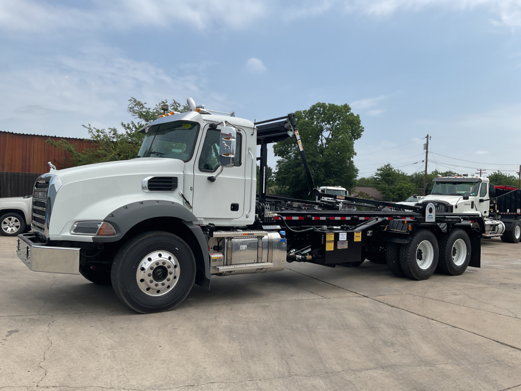Truck side view showing a white 2026 Mack GR64B roll-off with a heavy chassis and hook-lift system, front grille, round headlamps, and dual rear axles.