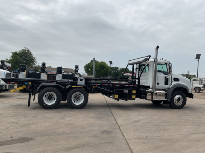 2026 Mack GR64B roll off truck side view showing cab, long chassis, and triple rear axles with a flatbed bed and hydraulic components.