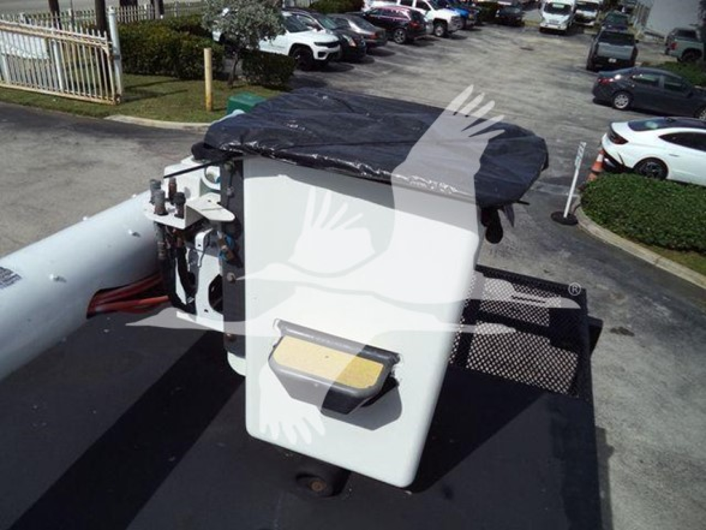 Bucket truck crane controls and white panel with hydraulic lines on top of truck bed, with black tarp covering and surrounding parking lot.