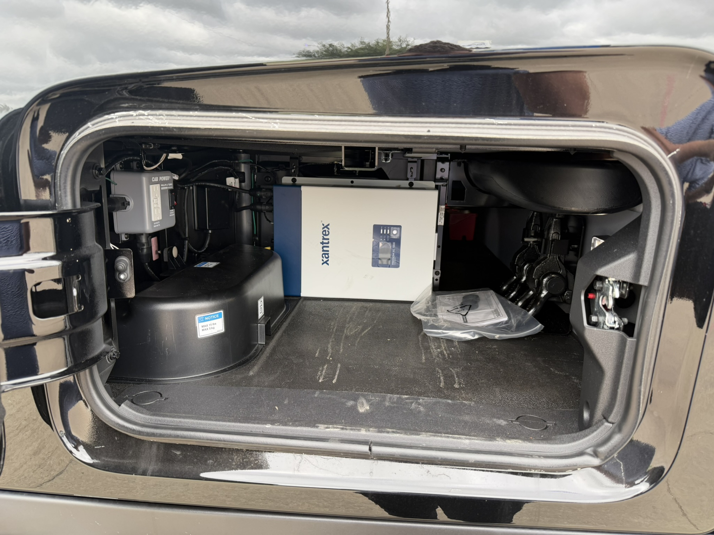 Front compartment of a 2027 Mack PR64T76H sleeper tractor showing a white Xantrex inverter box and other equipment in the storage bay.